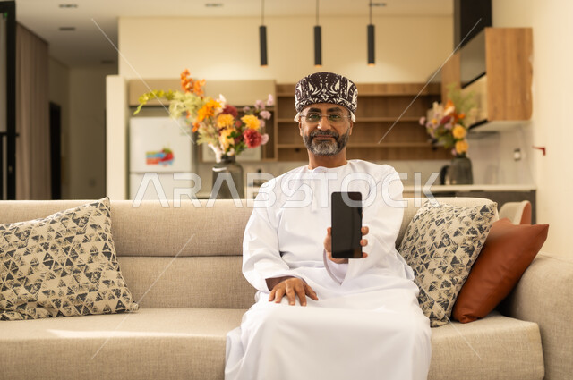 رجل عربي خليجي عماني يرتدي الدشداشة والكمة  يجلس على اريكة مريحة ويقوم بعرض شاشة سوداء فارغة عبر الهاتف المحمول، النظر الى الكاميرا بايماءات الفرح ، استخدام الأجهزة والتقنيات الحديثة والمتطورة .