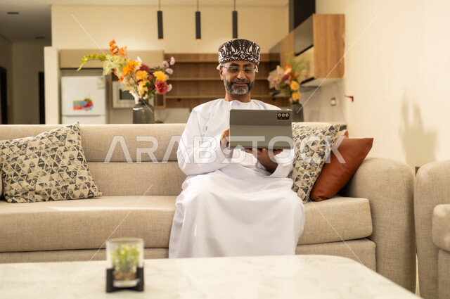 Knowing the latest news and browsing social media, tablet applications and programs, using modern technologies in remote work, an Arab Gulf Omani man wearing a dishdasha and kameh holding a tablet, completing work quickly and easily, integrating technology into daily life.