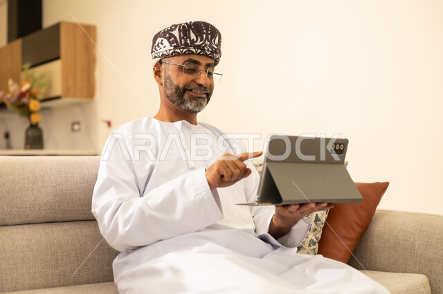 تطبيقات وبرامج الأجهزة اللوحية ، التطنور التقني والتكنولوجي ، انجاز مهام العمل عبر الانترنت ، رجل عربي خليجي عماني يرتدي الدشداشة والكمه يحمل بيده جهاز التابلت وينقرعليه باصبع السبابة، استخدام التقنيات الحديثة ، دمج التكنولوجيا في الحياة اليومية .