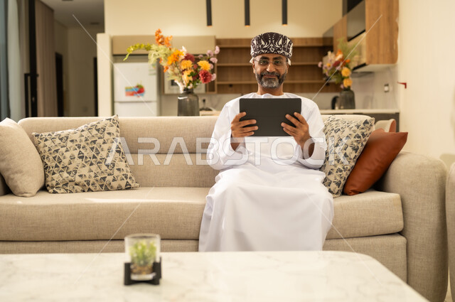 مفهوم إدارة المشاريع عن بعد، صورة لرجل عربي خليجي عماني يرتدي الدشداشة والكمة ويجلس على اريكة ويقوم بانجاز مهام العمل عبر جهاز التابلت، استخدام الاجهزة التقنية الحديثة ، تصفح مواقع ووسائل التواصل الاجتماعي .
