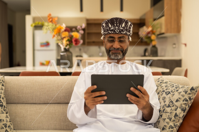 Holding online meetings, tablet applications and programs, using modern technologies in remote work, an Arab Gulf Omani man wearing a dishdasha and kameh holding a tablet, completing work quickly and easily.