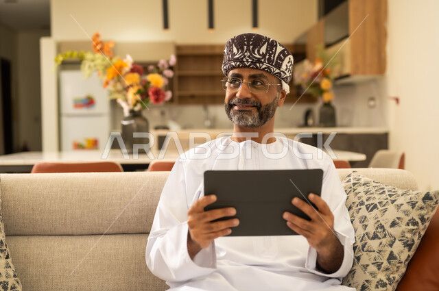 دمج التكنولوجيا في الحياة اليومية ، تطبيقات وبرامج الأجهزة اللوحية ، استخدام التقنيات الحديثة في العمل عن بعد، رجل عربي خليجي عماني يرتدي الدشداشة والكمه يحمل بيده جهاز التابلت وينظر الى شئ ما ، إنجاز الأعمال بسرعة وسهولة .