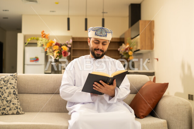 مفهوم التثقيف والبحث العلمي، القراءة بتمعن وتركيز، هواية المطالعة والاستكشاف، رجل عربي خليجي عماني يرتدي الدشداشة والكمة  يجلس على اريكة مريحة ويحمل بيده كتاب تثقيفي ،  الاستمتاع بقراءة الروايات والقصص .