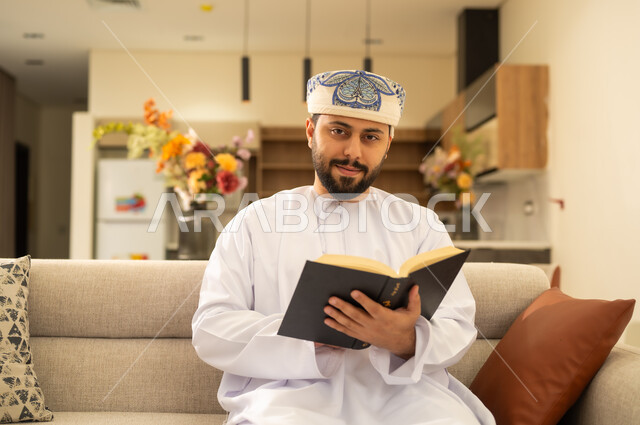 الاهتمام بتصفح الكتب المفيدة ، مفهوم التثقيف والبحث العلمي، القراءة بتمعن وتركيز، رجل عربي خليجي عماني يرتدي الدشداشة والكمة يحمل بيده كتاب تثقيفي وينظر الى الكاميرا ،  الاستمتاع بقراءة الروايات والقصص .