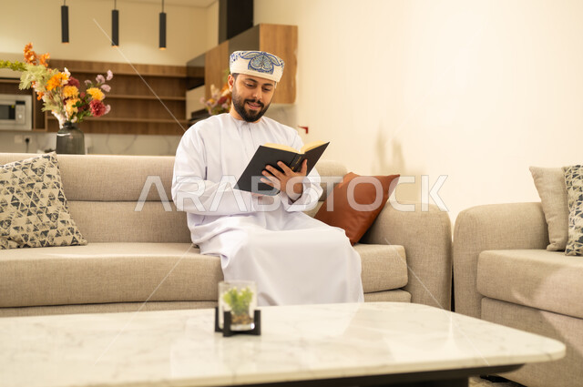هواية المطالعة والاستكشاف وقضاء وقت الفراغ بالتعلم والثقافة، رجل عربي خليجي عماني يرتدي الدشداشة والكمة  يجلس على اريكة مريحة ويحمل بيده كتاب تثقيفي ، القراءة بتمعن وتركيز، الاستمتاع بقراءة الروايات والقصص .