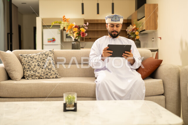 Tablet applications and programs, gestures of focus and seriousness, using modern technology and techniques, the concept of project management and completing tasks remotely. An Arab Gulf Omani man wearing a dishdasha and a kummah, holding a tablet, browsing websites and social media.