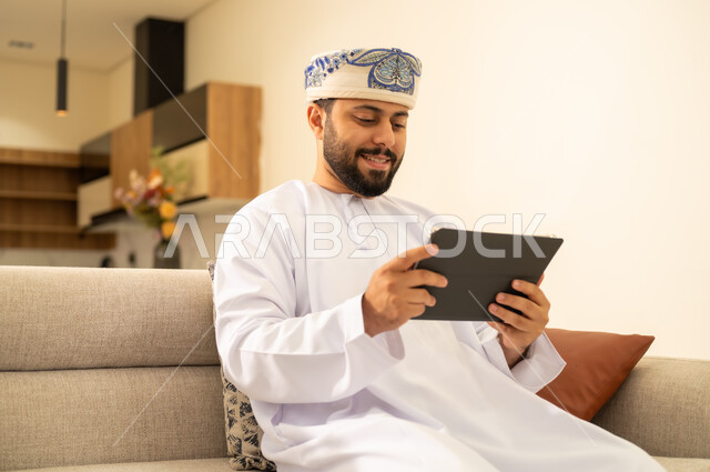 Watching movies and series online, using modern technology and techniques, tablet applications and programs, the concept of project management and completing tasks remotely. A close-up image of an Arab Gulf Omani man wearing a dishdasha and a kummah, holding a tablet, browsing websites and social media.