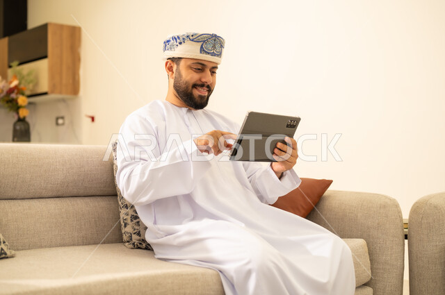 Using modern technologies in remote work; Completing tasks quickly and easily; An Arab Gulf Omani man wearing a dishdasha and a kimmah holding a tablet; tapping the device with his index finger; Tablet applications and programs.