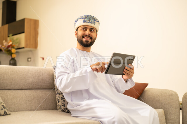 Looking at something with happy gestures, tapping the index finger on a tablet, using a modern technology device, completing tasks remotely, integrating technology into daily life, an Arab Gulf Omani man wearing a dishdasha and a keffiyeh holding a tablet, tablet applications and programs.