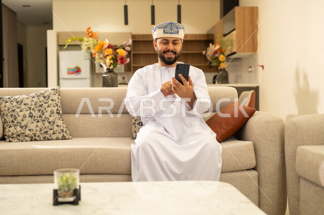 النظر إلى الجوال ومتابعة آخر الاخبار بإيماءات السعادة, استخدام جهاز تقني حديث ،  انجاز المهام عن بعد، دمج التكنولوجيا في الحياة اليومية ، رجل عربي خليجي عماني يرتدي الدشداشة والكمه ويحمل بيده هاتف محمول ، تصفح مواقع و وسائل التواصل الاجتماعي .
