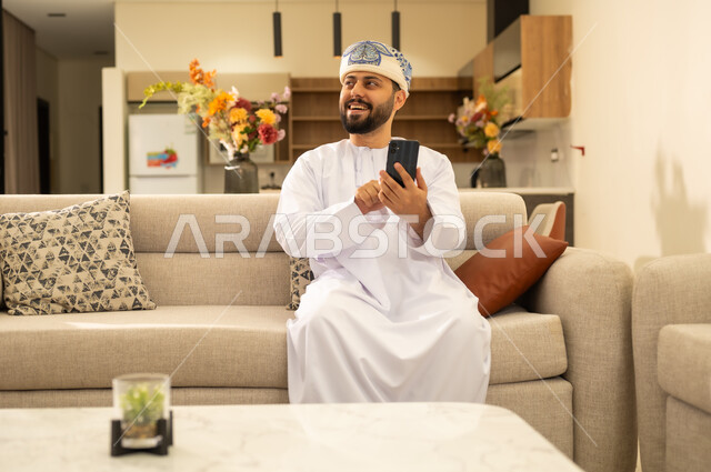 تصفح تطبيقات الهاتف المحمول, استخدام جهاز تقني حديث ، انجاز المهام عن بعد، دمج التكنولوجيا في الحياة اليومية ، رجل عربي خليجي عماني يرتدي الدشداشة والكمه يحمل بيده هاتف محمول وينظر الى شئ ما بايماءات السعادة ، تصفح مواقع و وسائل التواصل الاجتماعي .