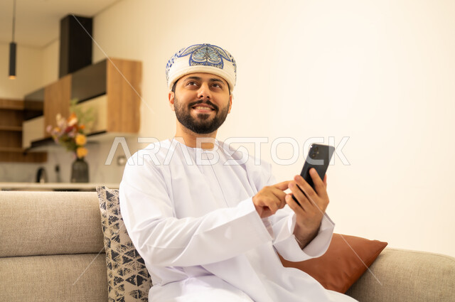 استخدام جهاز تقني حديث ،  انجاز المهام عن بعد، دمج التكنولوجيا في الحياة اليومية ، رجل عربي خليجي عماني يرتدي الدشداشة والكمه يحمل بيده هاتف محمول وينظر الى شئ ما بايماءات السعادة ، تصفح مواقع و وسائل التواصل الاجتماعي .