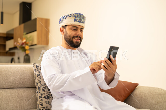 تصفح مواقع و وسائل التواصل الاجتماعي, انجاز المهام عن بعد، دمج التكنولوجيا في الحياة اليومية ، رجل عربي خليجي عماني يرتدي الدشداشة والكمه يجلس على اريكة مريحة ويقوم بإجراء مكالمة هاتفية عبر الجوال، استخدام الأجهزة الحديثة والمتطورة .