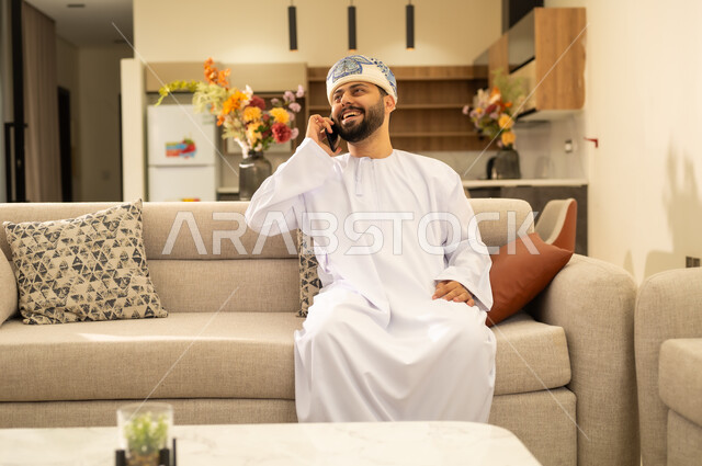 الرد على الاتصالات والمكالمات, صورة مقربة لرجل عربي خليجي عماني يرتدي الدشداشة والكمه يجلس على اريكة مريحة ويقوم بإجراء مكالمة هاتفية عبر الجوال، التواصل مع الأهل والأصدقاء عبر الهاتف المحمول، استخدام الأجهزة الحديثة والمتطورة.