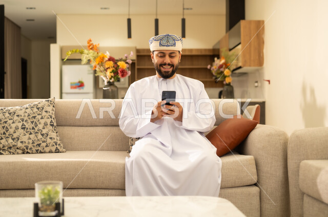 Completing tasks remotely, integrating technology into daily life. An Arab Gulf Omani man wearing a dishdasha and a kummah, holding a mobile phone, browsing social media, chatting with family and friends online.