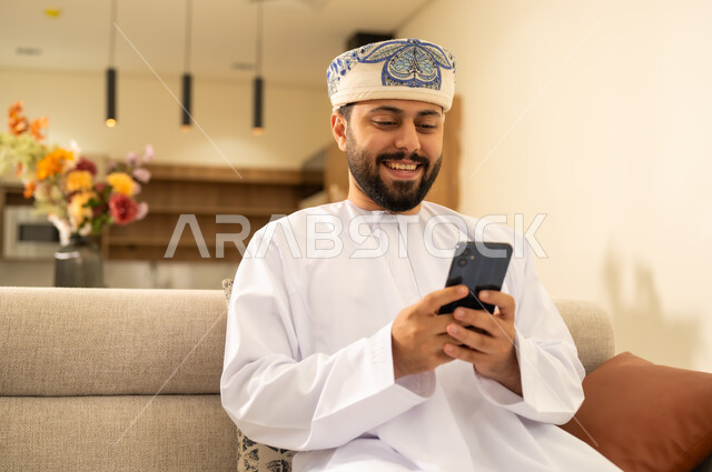 Integrating technology into daily life, using modern and advanced devices. An Arab Gulf Omani man wearing a dishdasha and a kummah, holding a mobile phone, browsing social media, chatting with family and friends online.