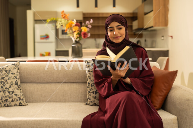 القراءة بتمعن وتركيز، قضاء وقت الفراغ بالتعلم والثقافة، إمراة عربية خليجية سعودية ترتدي العباءة والحجاب ، تجلس على اريكة مريحة وتحمل بيدها كتاب تثقيفي، امرأة عربية خليجية عمانية تفضل الاستمتاع بقراءة الروايات والقصص .