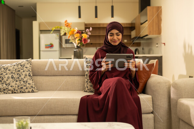 Browsing social media sites and platforms, integrating technology into daily life and work, a Saudi Arabian Gulf woman wearing an abaya and hijab sitting on a comfortable sofa, completing work tasks via a tablet, an Omani Arabian Gulf woman using modern technology devices, the concept of completing projects remotely.