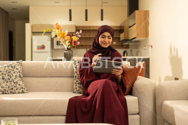 Remote project management concept, close-up of a Saudi Arabian Gulf woman wearing an abaya and hijab sitting on a sofa, completing work tasks via a tablet, an Omani Arabian Gulf woman browsing social media sites, using modern technical devices.