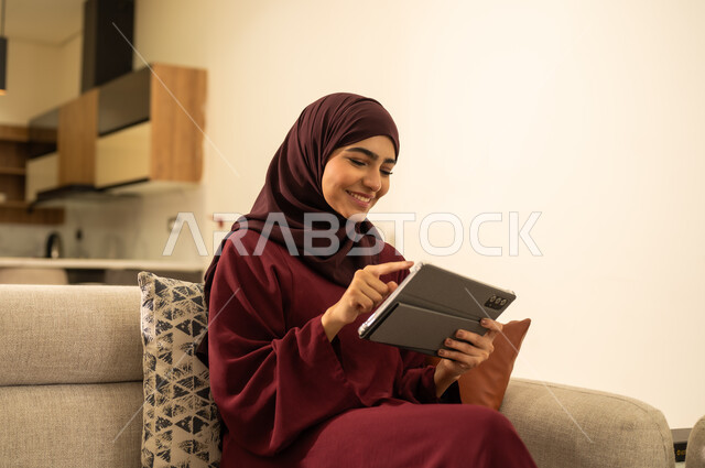 دمج التقنية في الحياة اليومية والعملية، صورة مقربة لامرأة عربية خليجية سعودية ترتدي العباءة والحجاب تجلس على اريكة مريحة ، تقوم بانجاز مهام العمل عبر جهاز التابلت، امرأة عربية خليجية عمانية تقوم باستخدام الاجهزة التقنية الحديثة ، تصفح مواقع ووسائل التواصل الاجتماعي، مفهوم انجار المشاريع عن بعد .