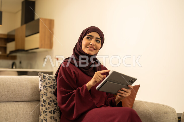الدردشة مع الأهل والأصدقاء عبر الانترنت، استخدام التكنولوجيا و التقنيات الحديثة, تطبيقات وبرامج الأجهزة اللوحية، صورة مقربة لامرأة عربية خليجية سعودية ترتدي العباءة والحجاب وتجلس على اريكة مريحة وتنظر إلى شيء ما بإيماءات السرور والسعادة، ، امرأة عربية خليجية عمانية تقوم بانجاز مهام العمل عبر جهاز التابلت، تصفح مواقع ووسائل التواصل الاجتماعي .