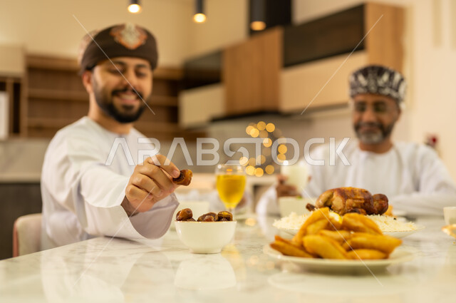 مائدة افطار رمضانية ، ايماءات السعادة بقدوم الشهر الفضيل ، شاب عربي خليجي عماني يرتدي الدشداشة والكمة و يتناول حلوي اللقيمات ، اجتماع الأهل ومشاركة الطعام على مائده الإفطار ، اجواء وزينة رمضانية ، المقبلات والمأكولات العربية الشرقية ، شهر الخير والبركة .