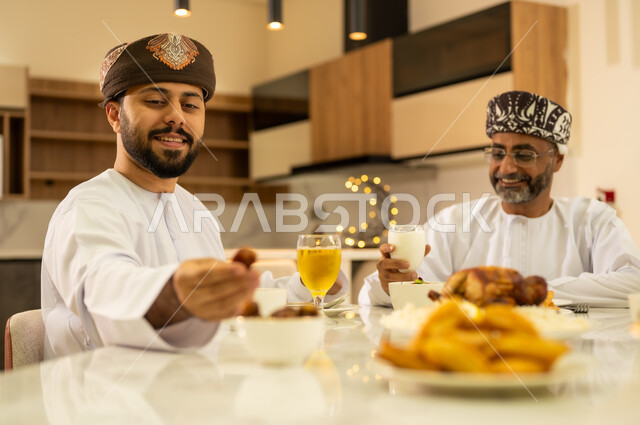 مفهوم الترابط و الأجواء الرمضانية العائلية ، سفرة مليئة باصناف من الأطعمة والمقبلات الشهية ، إجتماع الاسرة في وقت الإفطار ، شاب عربي خليجي عماني يرتدي الدشداشة والكمة و يتناول حبات التمر ، ايماءات وجه سعيدة ، اجواء وزينة رمضانية ، شهر الخير والبركة .