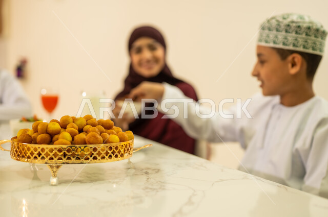 صورة مقربة لحلوى اللقيمات الشهية ، العزائم والولائم فى الاعياد والمناسبات الدينية الاسلامية ، اجواء عائلية دافئة بايماءات وجه سعيدة ، عائلة عربية خليجية عمانية تجتمع على مائدة الافطار فى شهر رمضان, المقبلات والمأكولات العربية الشرقية  ، ديكورات وزينة رمضانية ، شهر الخير والبركة .