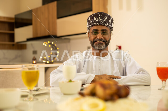 Spending quality time with family at the iftar table. An Arab Gulf Omani man wearing a dishdasha and a kummah looks at the camera with happy gestures. Celebrating the arrival of the holy month. Delicious Ramadan foods and appetizers. Decorations and décor for the month of goodness and blessing.s