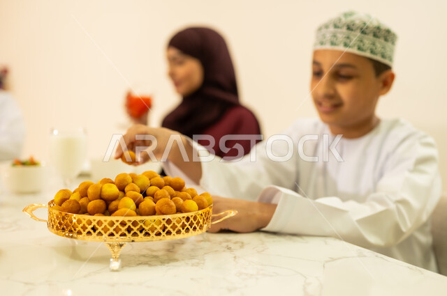 ايماءات السعادة بقدوم الشهر الفضيل ، ولد عربي خليجي عماني يرتدي الدشداشة والكمة و يتناول حلوي اللقيمات ، ايماءات وجه سعيدة ، اجتماع الأهل ومشاركة الطعام على مائده الإفطار ، اجواء وزينة رمضانية ، المقبلات والمأكولات العربية الشرقية ، شهر الخير والبركة .