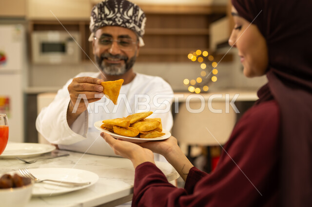 تقدير واحترام المرأه لزوجها ، اجتماع الأهل ومشاركة الطعام على مائده الإفطار ، امراه عربية خليجية عمانية تقوم بتقديم طبق من السمبوسك المقرمشة لزوجها ، مقبلات رمضانية شهية، فطائر مقلية متنوعة الحشوات,  اجواء عائلية دافئة بايماءات وجه سعيدة  ، الاحتفال بقدوم الشهر الكريم ، ديكورات وزينة رمضانية ، شهر الخير والبركة .