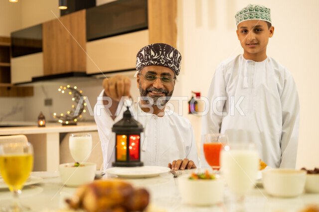 الاحتفال بقدوم شهر رمضان المبارك ، اضاءة وزينة شهر الخير والبركة ، مائدة افطار مليئة بأصناف من المأكولات والمقبلات العربية، أب عربي خليجي عماني مبتسم يرتدي الدشداشه و الكمه يمسك بيده فانوس معدني مضيء ، مناسبات دينية اسلامية .