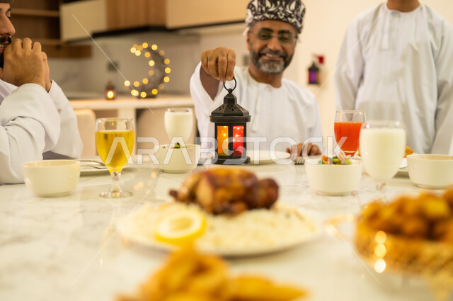 مائدة الافطار  فى شهر رمضان ، سفرة مليئة بأصناف من المأكولات والمقبلات العربية، أب عربي خليجي عماني مبتسم يرتدي الدشداشه و الكمه يمسك بيده فانوس معدني مضيء ، اضاءة وزينة شهر الخير والبركة ، مناسبات دينية اسلامية .
