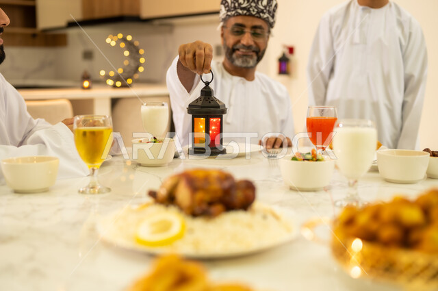 تناول الافطار الرمضاني عند أذان المغرب ، الاحتفال بقدوم شهر رمضان المبارك ، مائدة افطار مليئة بأصناف من المأكولات والمقبلات العربية، أب عربي خليجي عماني مبتسم يرتدي الدشداشه و الكمه يمسك بيده فانوس معدني مضيء ، مناسبات دينية اسلامية .