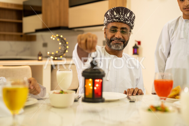 إضاءة وزينة شهر رمضان الكريم , صورة مقربة لرجل عربي خليجي عماني مبتسم يرتدي الدشداشه و الكمه يمسك بيده فانوس معدني مضيء ، مفهوم التجمعات العائلية السعيدة في شهر رمضان المبارك ، شهر الخير والبركة.