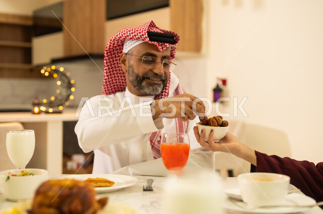 منتجات محلية سعودية ، أداء فريضة الصيام ، حسن الكرم والضيافة في المملكة العربية السعودية ، صورة مقربة ليد إمراه عربية خليجية سعودية تقدم طبق من التمر السعودي لزوجها على مائدة الافطار فى شهر رمضان, المقبلات والمأكولات الشرقية السعودية ، اجواء وزينة رمضانية ، شهر الخير والبركة .