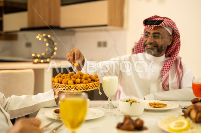 حلويات شرقية لذيذة ، سفرة مليئة باصناف من الأطعمة والمقبلات الشهية، الأجواء الرمضانية السعيدة ، إجتماع الاسرة في وقت الإفطار ، رجل عربي خليجي سعودي يرتدي الشماغ والزي التقليدي و يتناول حلوي اللقيمات ، ايماءات وجه سعيدة ، اجواء وزينة رمضانية ، شهر الخير والبركة .