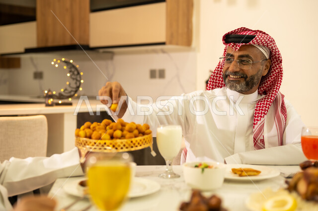 تناول الأطعمة والمقبلات الشهية، الأجواء الرمضانية السعيدة ، إجتماع الاسرة في وقت الإفطار  ، رجل عربي خليجي سعودي يتناول حلوي اللقيمات ، ايماءات وجه سعيدة ، اجواء وزينة رمضانية ، شهر الخير والبركة .