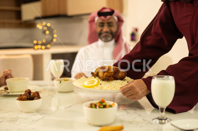تجهيز مائدة الافطار ، أم عربية خليجية سعودية تقوم بتحضير سفرة رمضان و وضع الطبق الرئيسي عليها, ايماءات وجه سعيدة  ، قدوم الشهر الكريم, الاحتفال بالاعياد و المناسبات الاسلامية, اجواء عائلية دافئة ، ديكورات وزينة رمضانية ، شهر الخير والبركة .