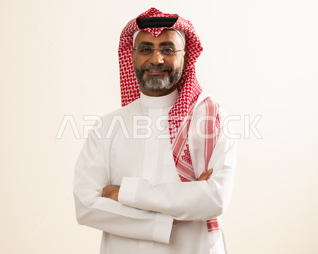 النظر الى الكاميرا  بإيماءات السرور والثقة بالنفس، مفهوم الرجولة والاناقة والاهتمام بالمظهر الخارجي ، صورة شخصية للوثائق الرسمية ، بورتريه مقرب لرجل عربي خليجي سعودي مبتسم يرتدي الشماغ والثوب التقليدي يقف بإستقامة وأيدي متقاطعة ، خلفية بيج.