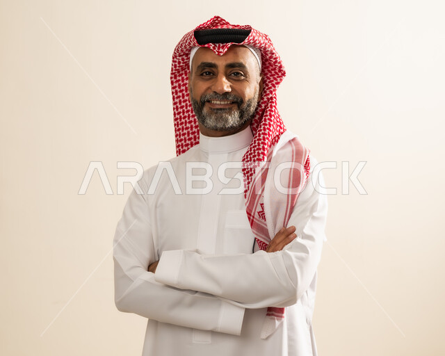 الوقوف بأيدي متقاطعة بتعابير الثقة بالنفس ، الاهتمام بالأناقة و المظهر الخارجي ، بورتريه مقرب لرجل عربي خليجي سعودي مبتسم يرتدي الشماغ والثوب التقليدي يقف بإستقامة و ينظر إلى الكاميرا بإيماءات البشاشة والسرور ، خلفية بيج.