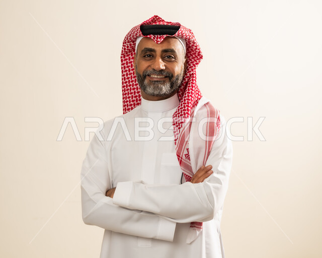 صورة شخصية للوثائق الرسمية، مفهوم الاهتمام بالمظهر الخارجي والرجولة والأناقة، بورتريه مقرب لرجل عربي خليجي سعودي مبتسم يرتدي الشماغ والثوب التقليدي يقف بإستقامة وأيدي متقاطعة ينظر إلى الكاميرا بإيماءات البشاشة والسرور، خلفية بيج.