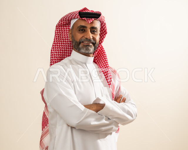 مفهوم الوسامة والرجولة والهيبة، الاهتمام بالأناقة و المظهر الخارجي ، بورتريه مقرب لرجل عربي خليجي سعودي مبتسم يرتدي الشماغ والثوب التقليدي يقف بإستقامة وأيدي متقاطعة ينظر إلى الكاميرا بإيماءات البشاشة والسرور والثقة بالنفس ، خلفية بيج.