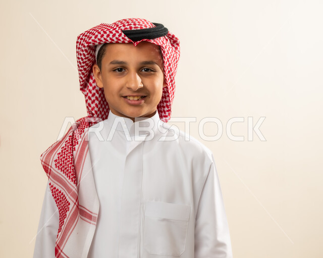 التقاط صورة شخصية للوثائق الرسمية ، بورتريه مقرب لولد عربي خليجي سعودي مبتسم يرتدي الشماغ والزي التقليدي وينظر الى الكاميرا بايماءات السرور، الوقوف باستقامة وثقة بالنفس، مفهوم الاهتمام بالمظهر الخارجي ، خلفية بيج .