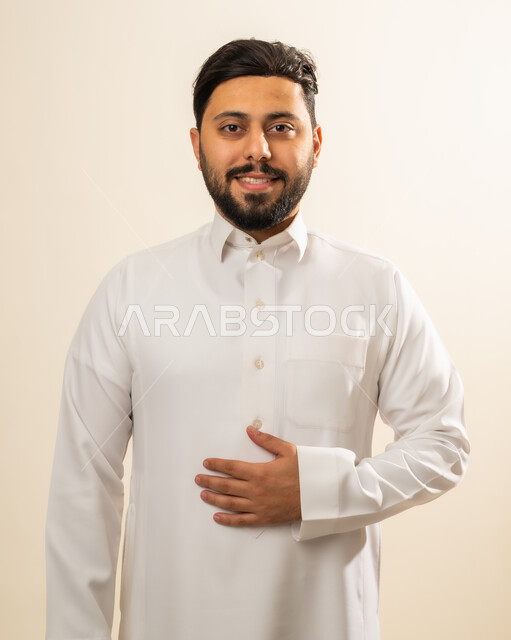 ايماءات الثقة والاعتزاز بالنفس، الاهتمام بالمظهر الخارجي، مفهوم الرجولة والأناقه، بورتريه لرجل عربي خليجي سعودي مبتسم يرتدي الزي التقليدي وينظر إلى الكاميرا بتعابير السرور، خلفية بيج .