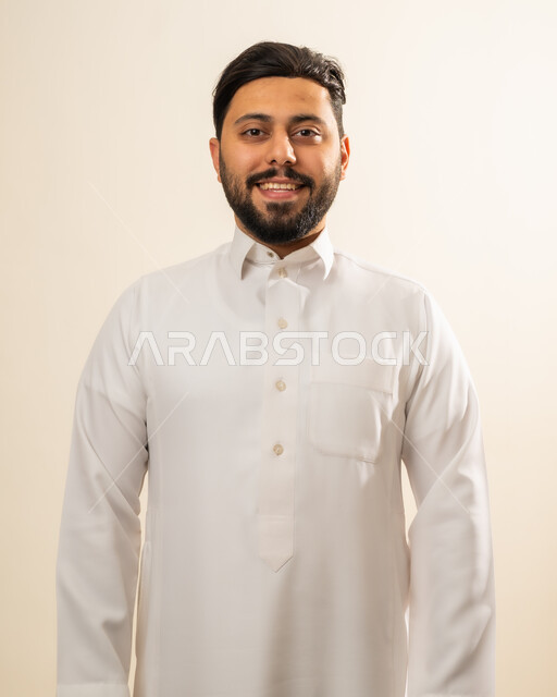 مفهوم الرجولة والقوة والثقة بالنفس، بورتريه لرجل عربي خليجي سعودي مبتسم يرتدي الزي التقليدي ينظر إلى الكاميرا بإيماءات السرور والفرح، الاهتمام والأناقه في المظهر الخارجي، خلفيه بيج.