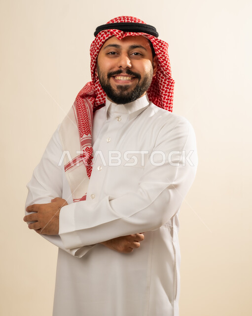 .The concept of elegance and masculinity, interest in outward appearance, portrait of a smiling Saudi Arabian Gulf man wearing a traditional shemagh and thobe, standing with crossed hands looking at the camera with an expression of self-confidence, beige background