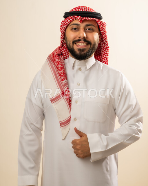تعابير الإعجاب والرضى، مفهوم القبول والموافقه، بورتريه لرجل عربي خليجي سعودي يرتدي الشماغ و الزي التقليدي يرفع اصبع الابهام بإيماءات الامتياز والجودة، خلفية بيج.
