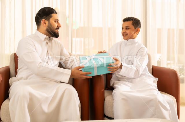 تبادل نظرات المحبة والألفة بين الأب والابن، إدخال السعادة والسرور إلى قلب الأهل, ولد عربي خليجي سعودي يرتدي الثوب التقليدي , يقدم هدية لوالده في أجواء العيد , إيماءات الفرح و البهجة, تقديم الهدايا فى المناسبات و العيد السعيد .