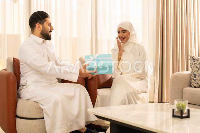 .The concept of intimacy and love between spouses, a Saudi Arabian Gulf man wearing traditional dress presents a gift to his wife with gestures of surprise and happiness, giving souvenirs on Mother's Day, sharing holidays, celebrations and happy occasions
